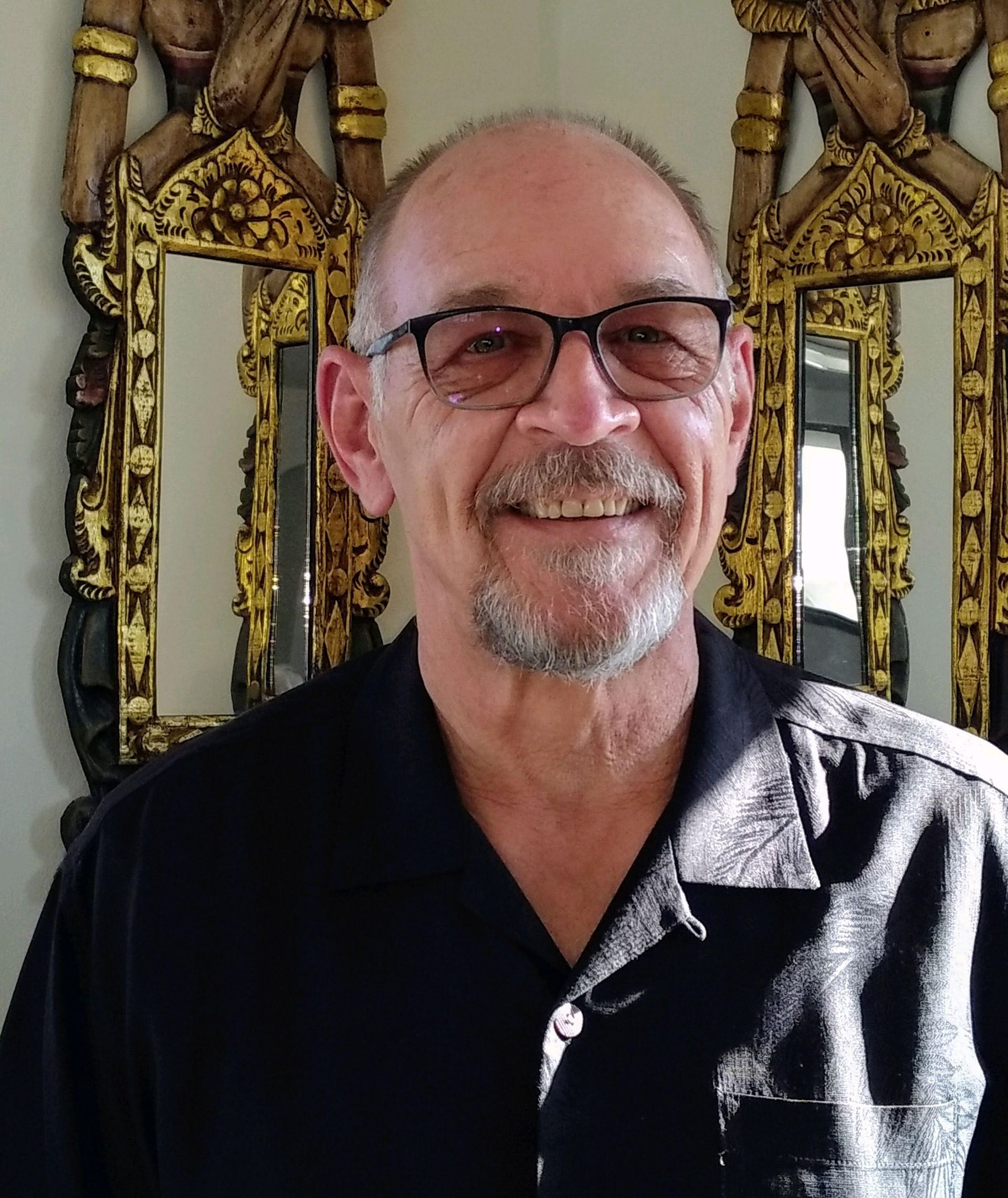 Smiling man in glasses and black shirt standing before ornate gold-framed mirrors