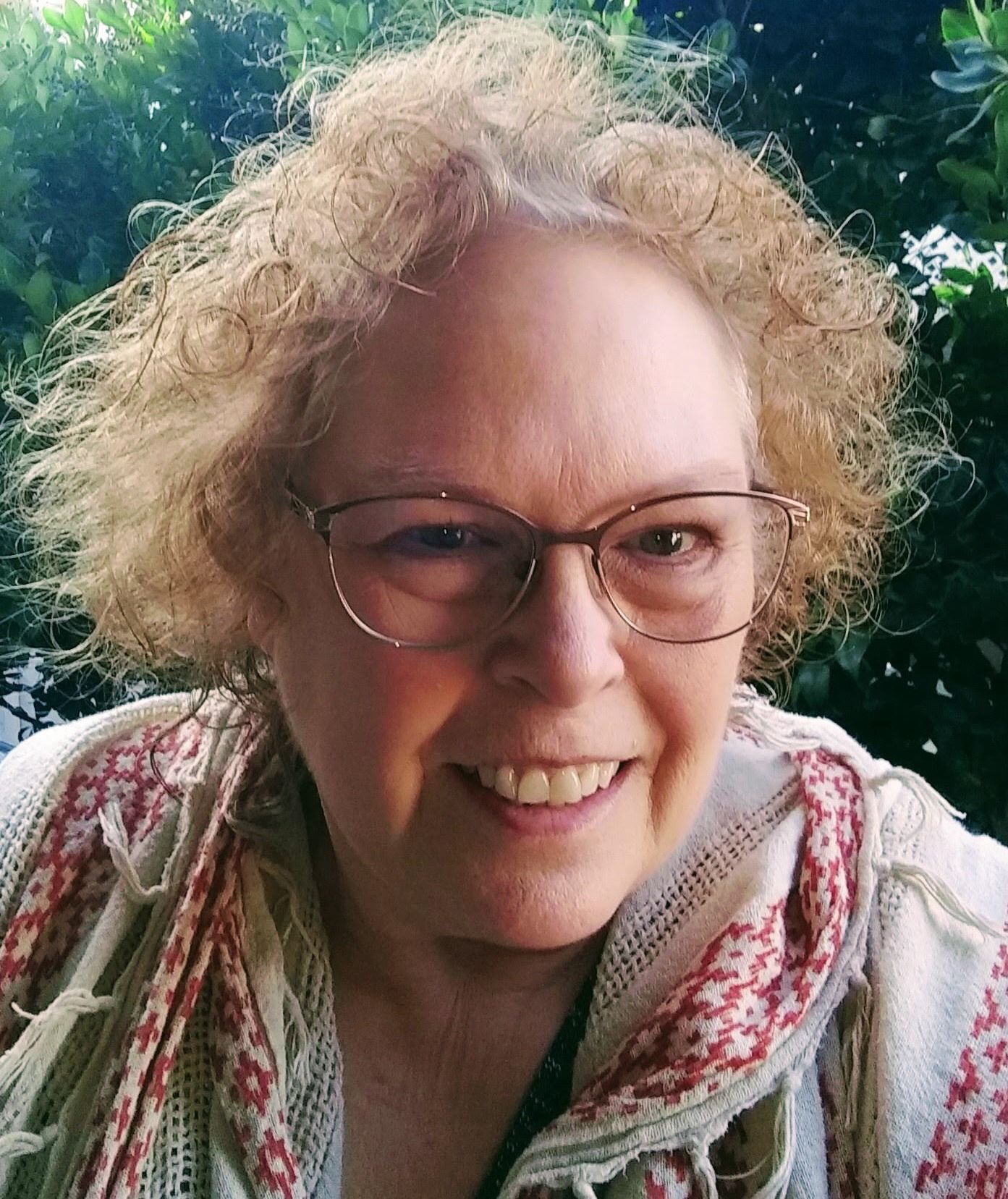 Smiling person with curly hair and glasses, wearing a patterned scarf, standing outdoors by greenery