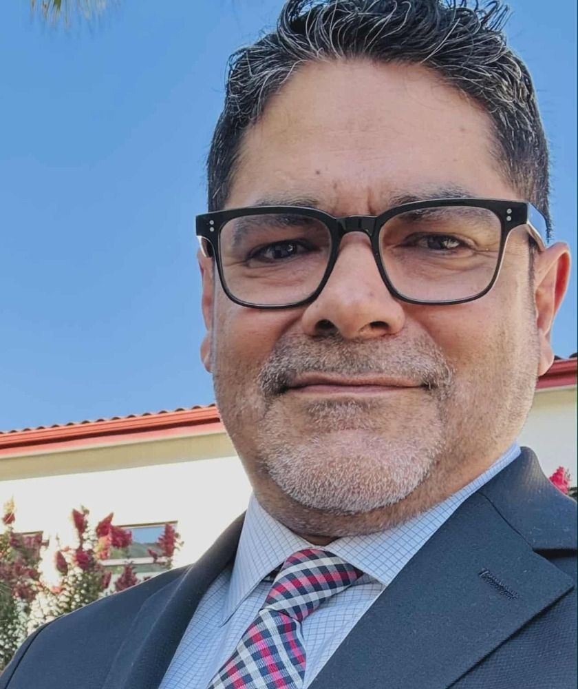 Man in glasses and suit smiling outdoors under a blue sky.