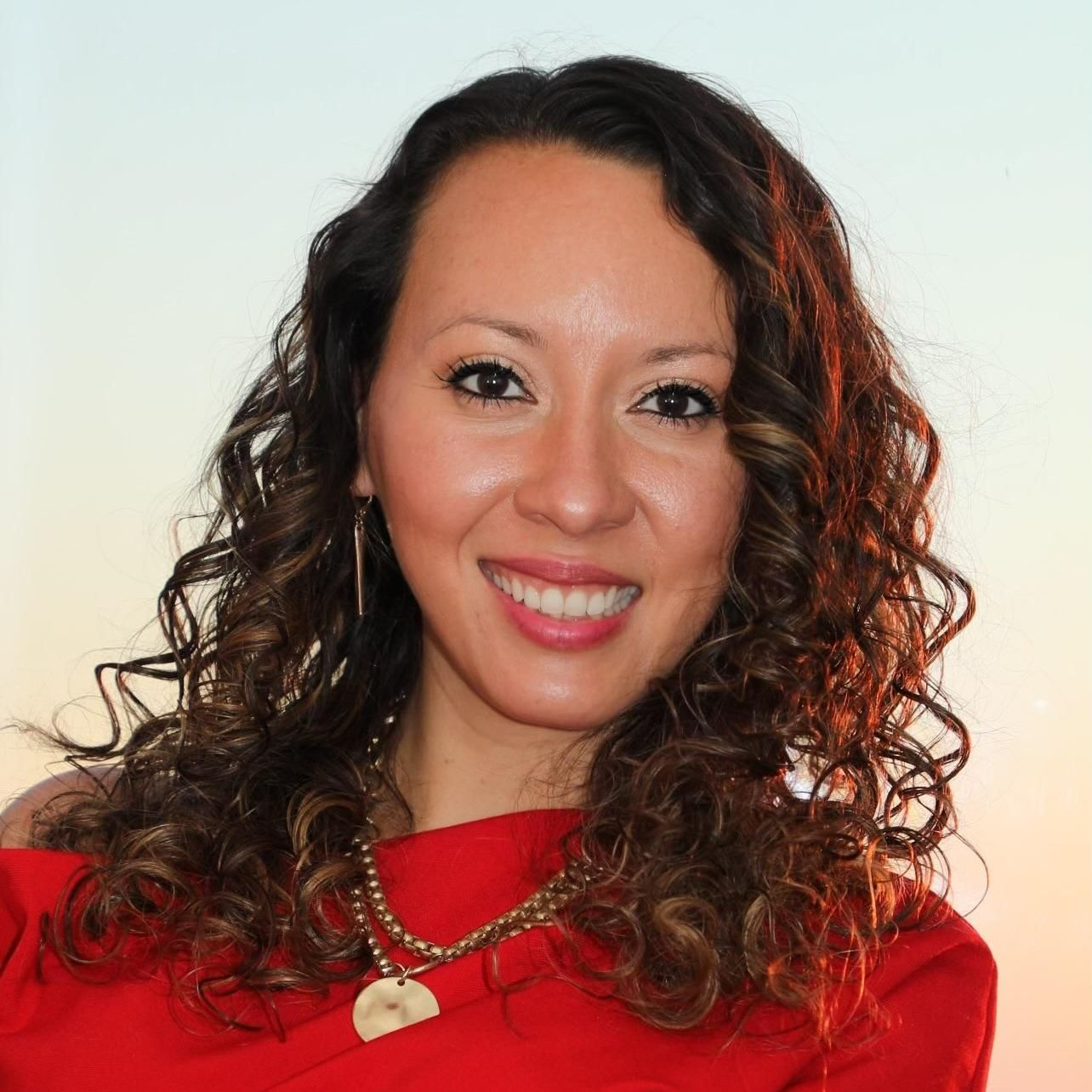 Woman with curly brown hair smiles in a red top with a necklace; soft sunset background.