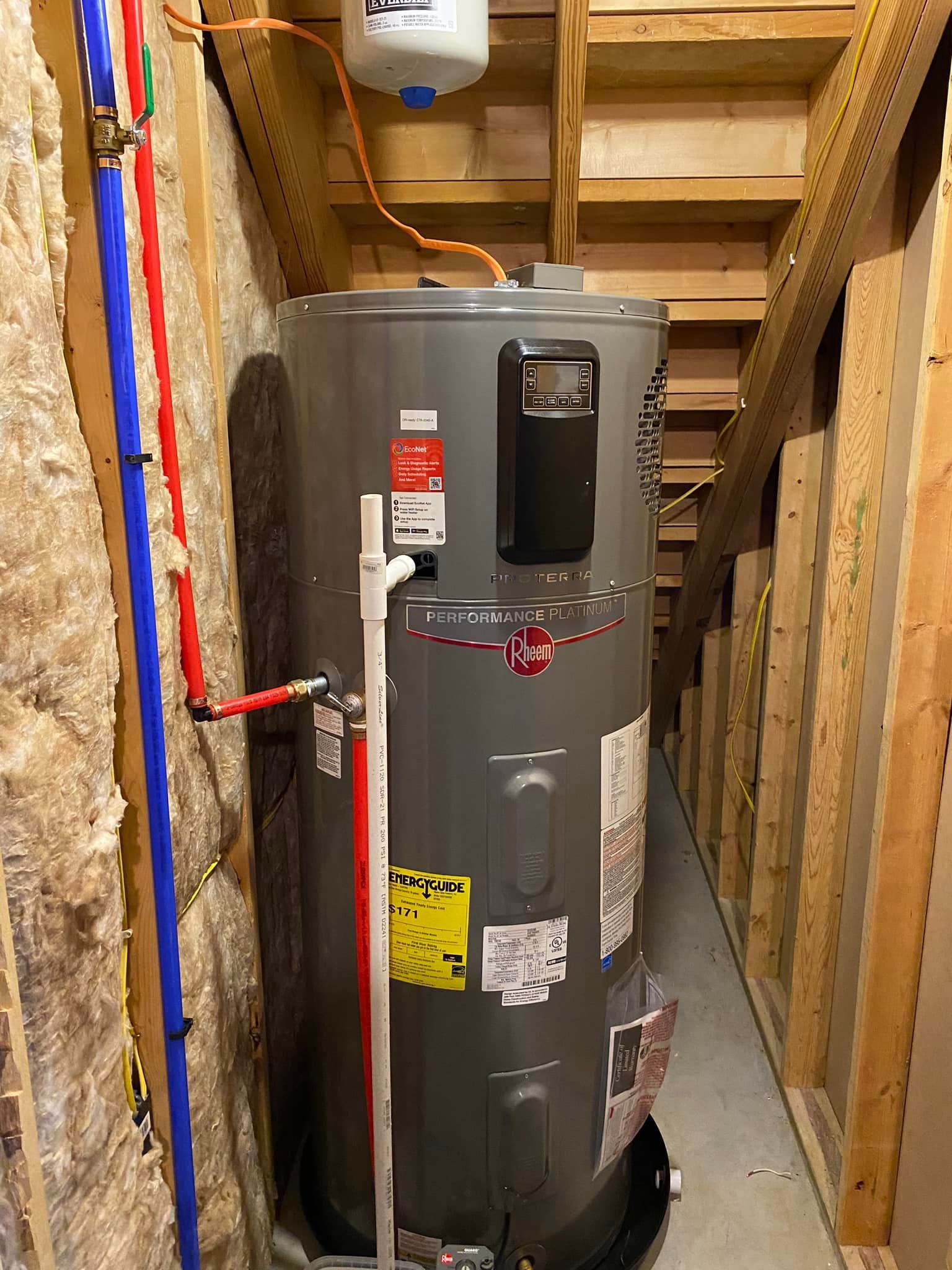 A gray electric water heater installed in an unfinished basement area with exposed insulation and wooden framing.