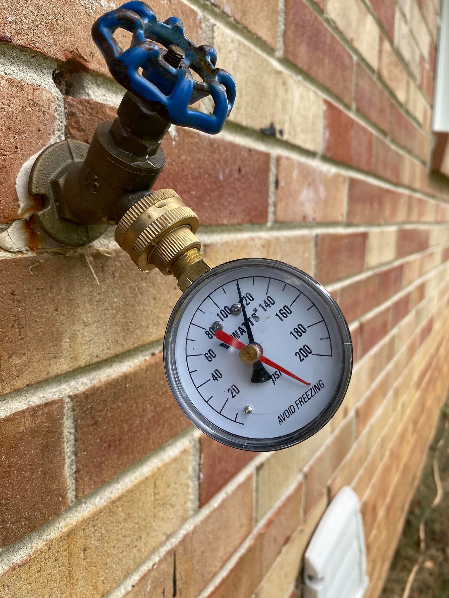 A pressure gauge attached to an outdoor brick wall water spigot with a blue handle, indicating a reading of 80 PSI.