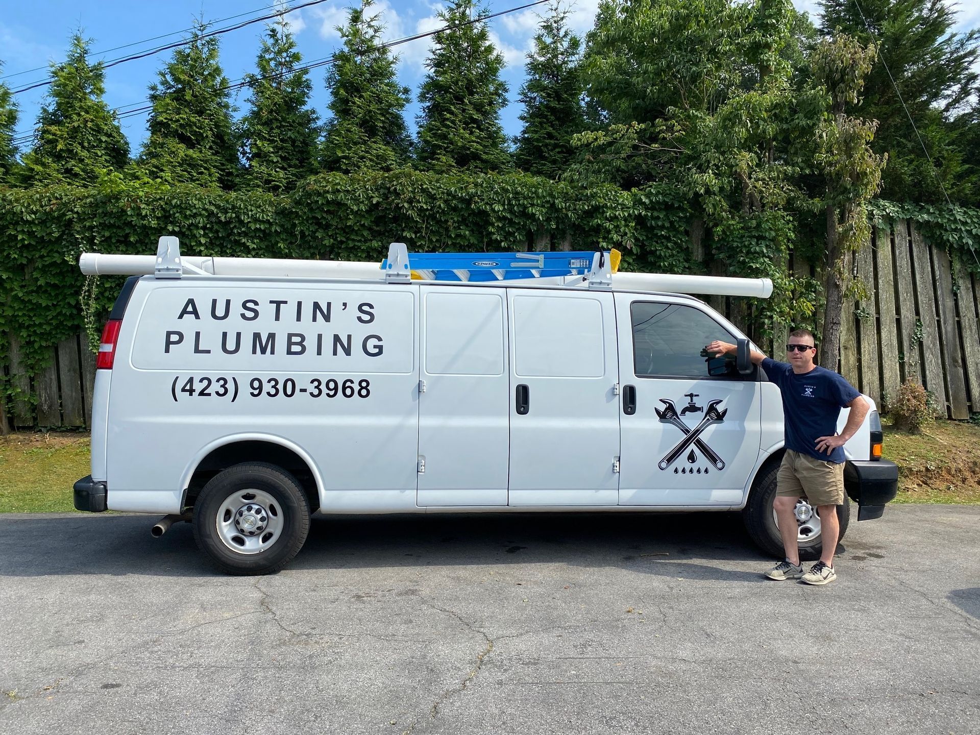 A person stands next to a white Austin's Plumbing van with a ladder rack and company logo, parked in front of a hedge.
