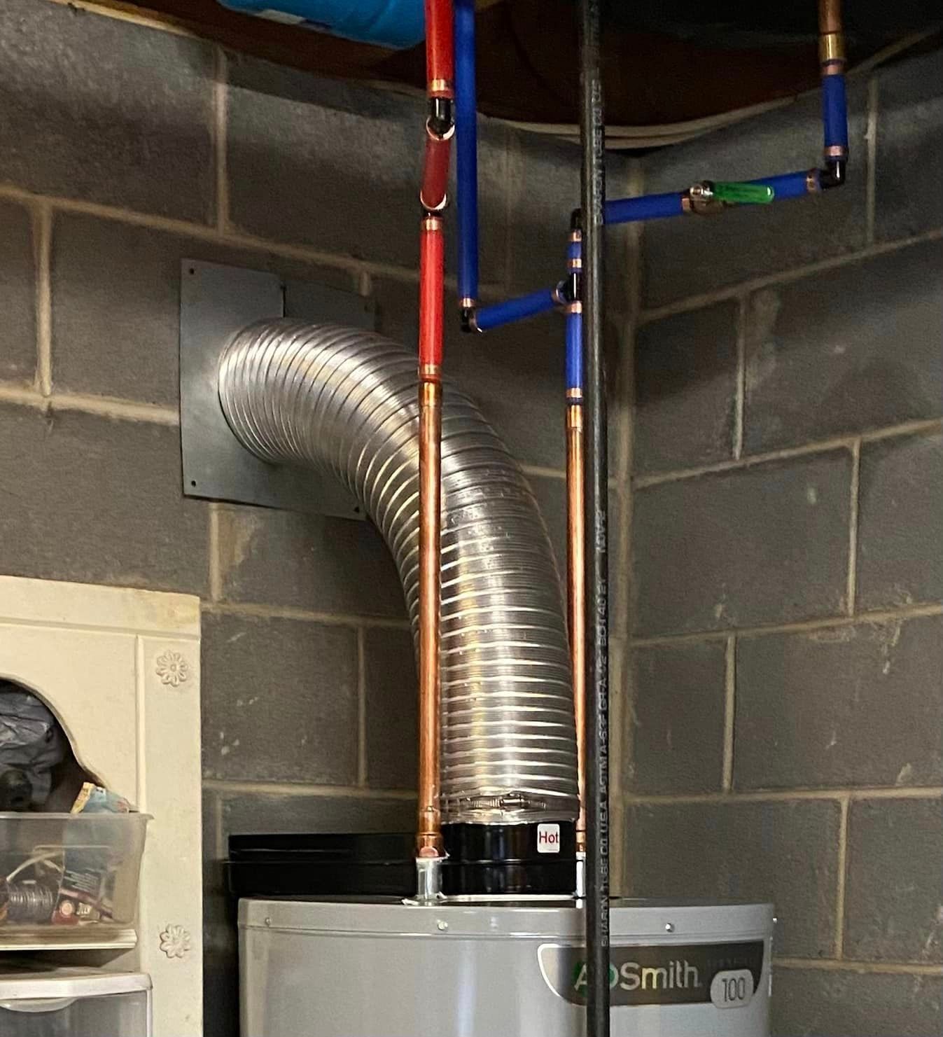 Water heater with metal exhaust venting and blue and red PEX plumbing pipes installed against a grey concrete block wall.