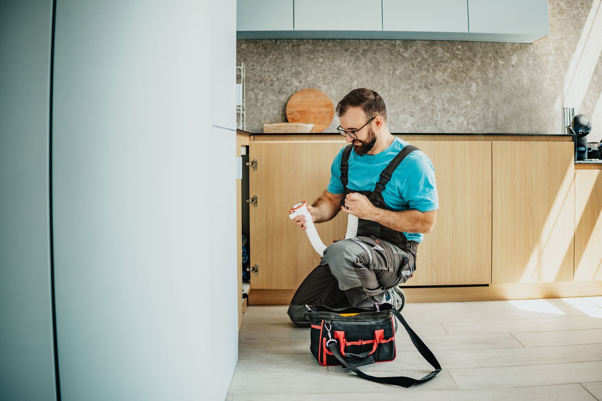 A person in a blue shirt and work overalls kneeling in a kitchen, holding a white plumbing pipe with a tool bag nearby.