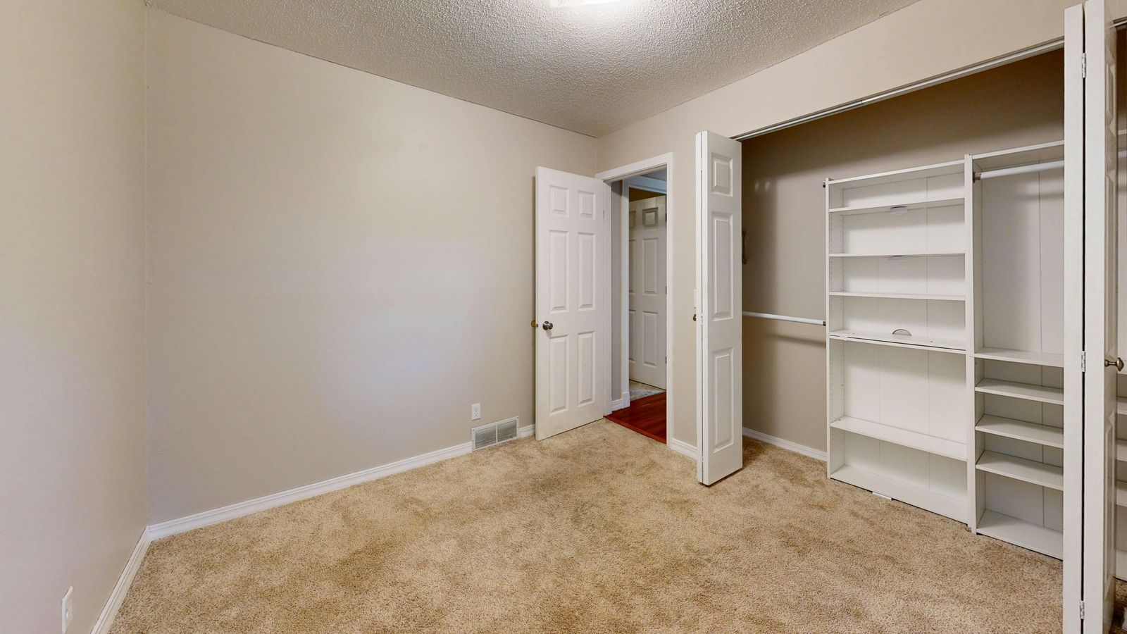 Empty beige bedroom with carpet, open closet shelving, and an open door to a hallway