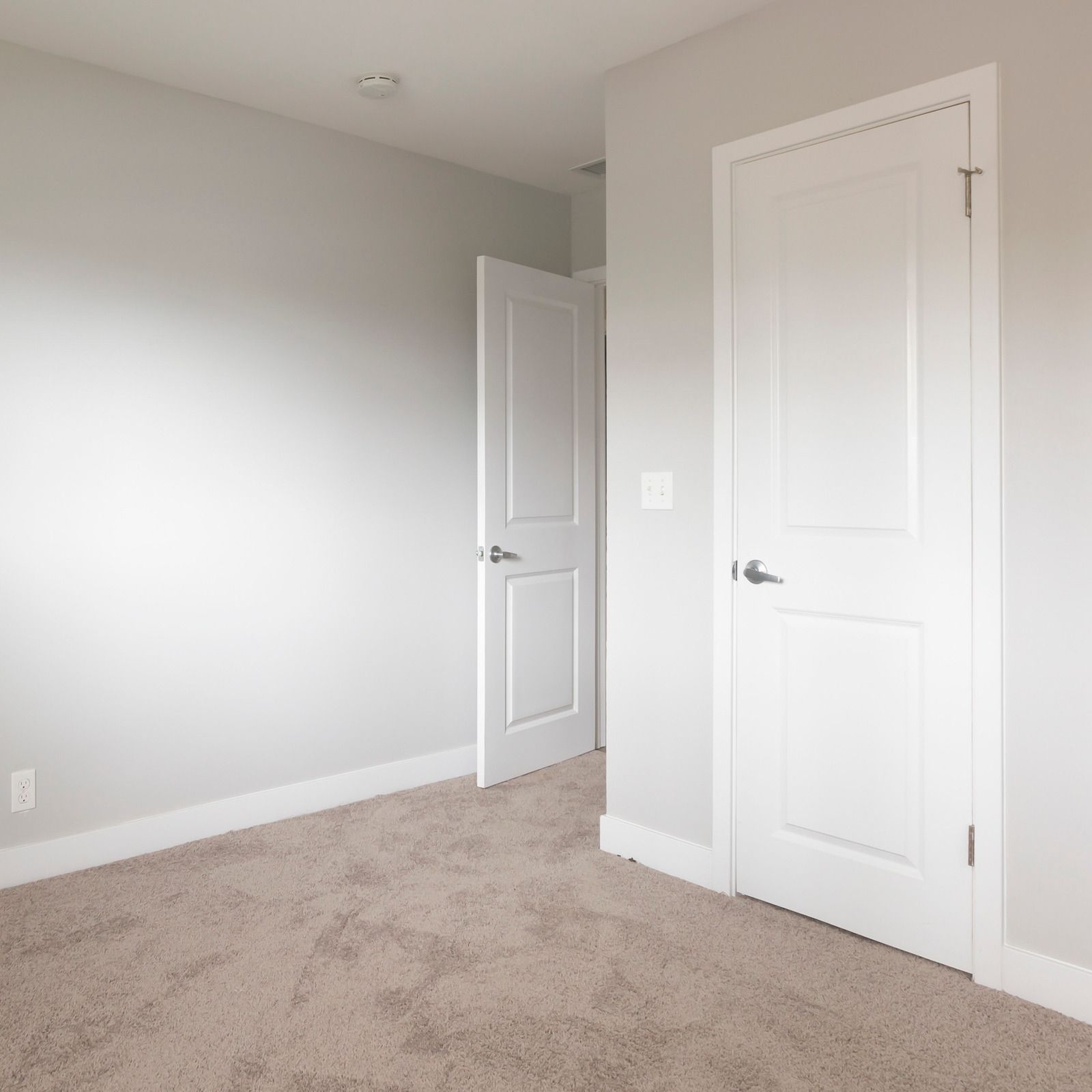 Empty bedroom with light gray walls, beige carpet, and two white doors, one partially open.