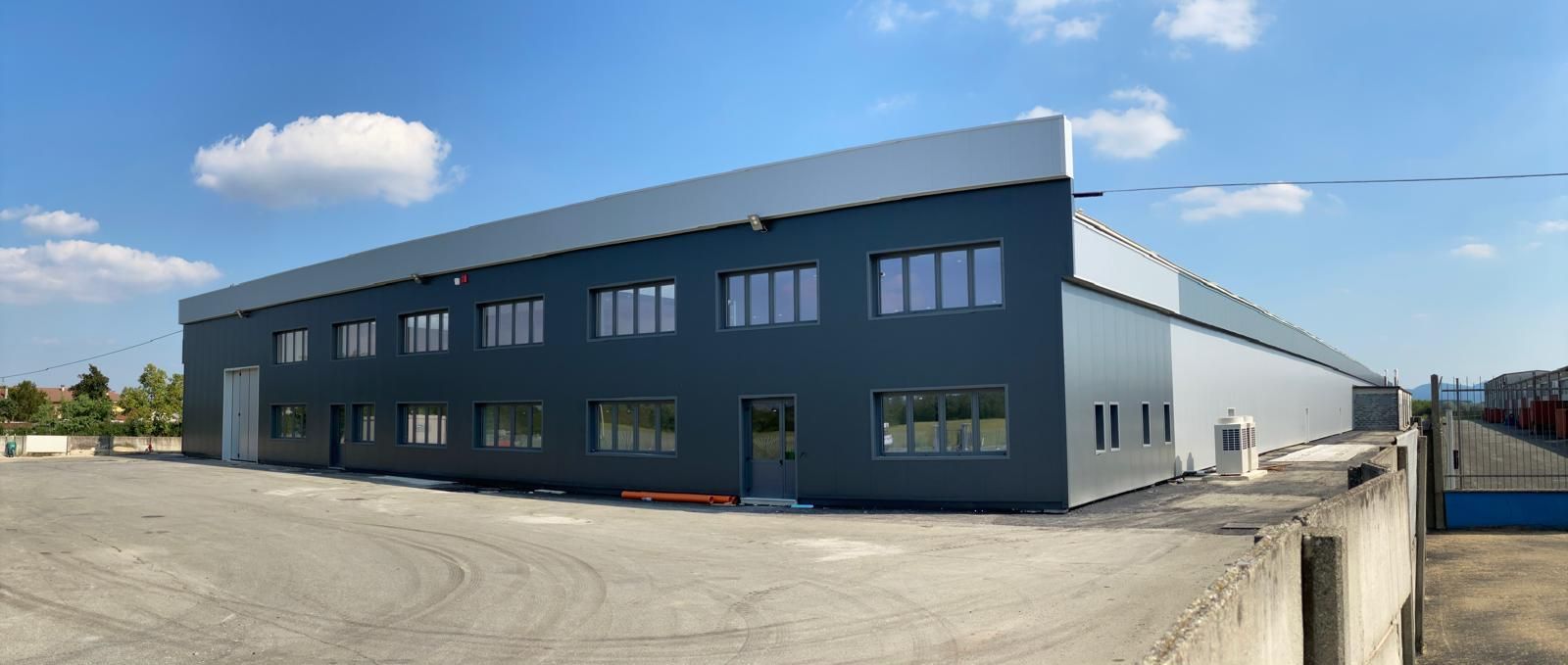Ampio magazzino industriale grigio con tetto bianco inclinato e finestre su un lotto pavimentato sotto un cielo azzurro.