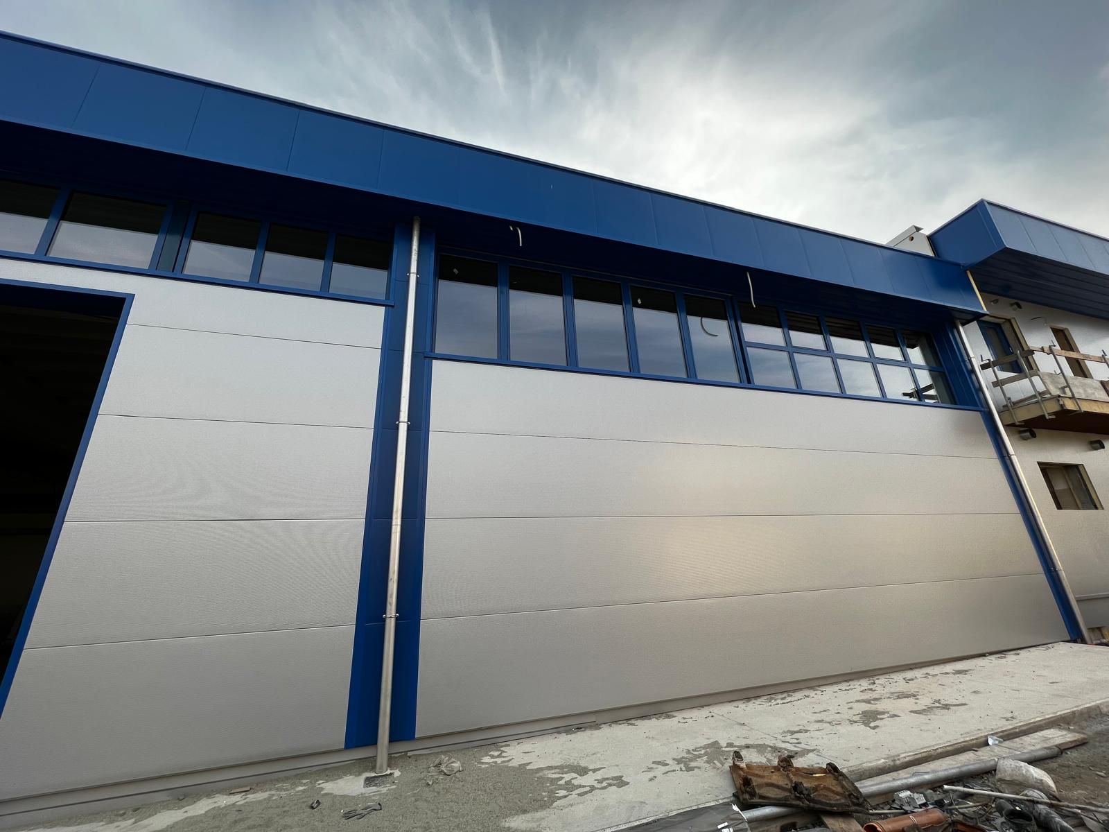Esterno di un edificio industriale blu e bianco con un grande portone in stile garage e finestre sotto un cielo nuvoloso.