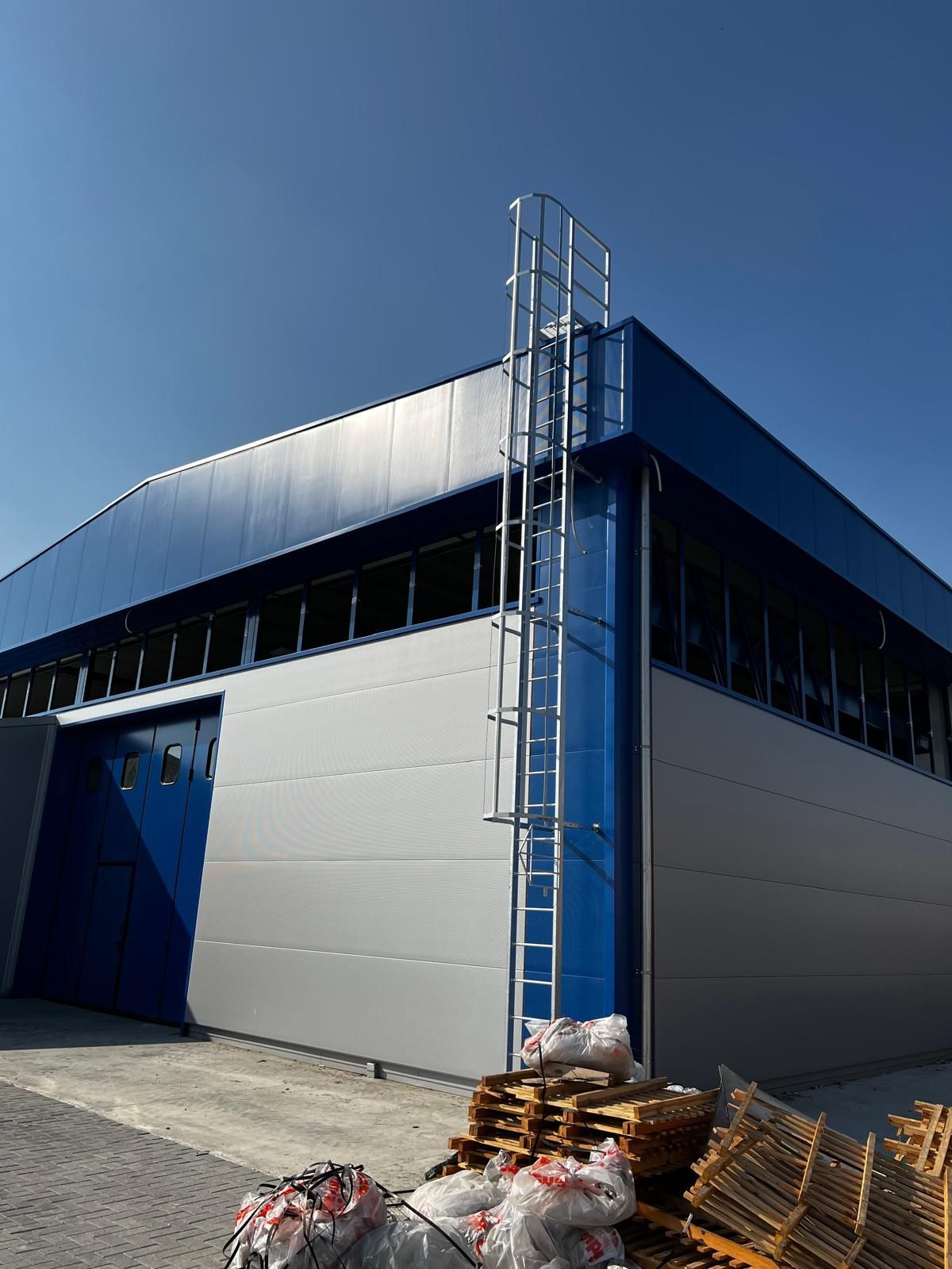 Moderno magazzino industriale con pannelli grigio-blu, una porta blu e materiali accatastati all'esterno sotto un cielo limpido.