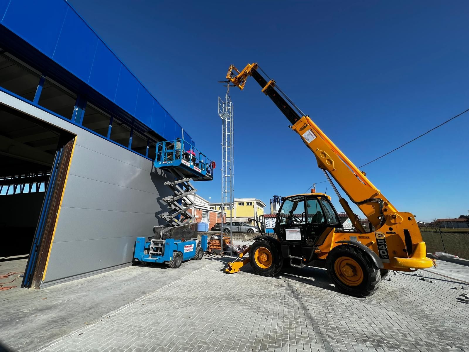 Cantiere edile con sollevatore telescopico per materiali accanto a un edificio industriale blu.