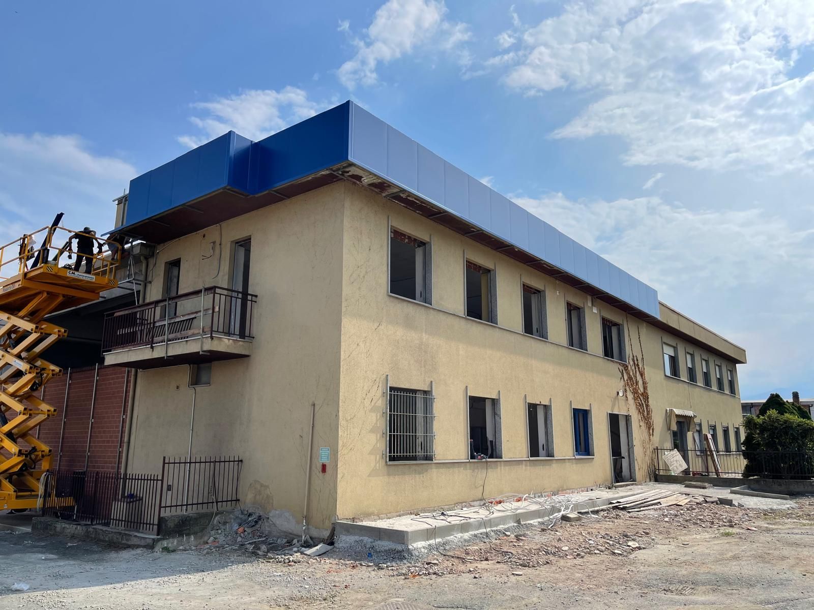 Edificio beige a due piani in costruzione con pannelli del tetto blu e impalcature sulla sinistra.