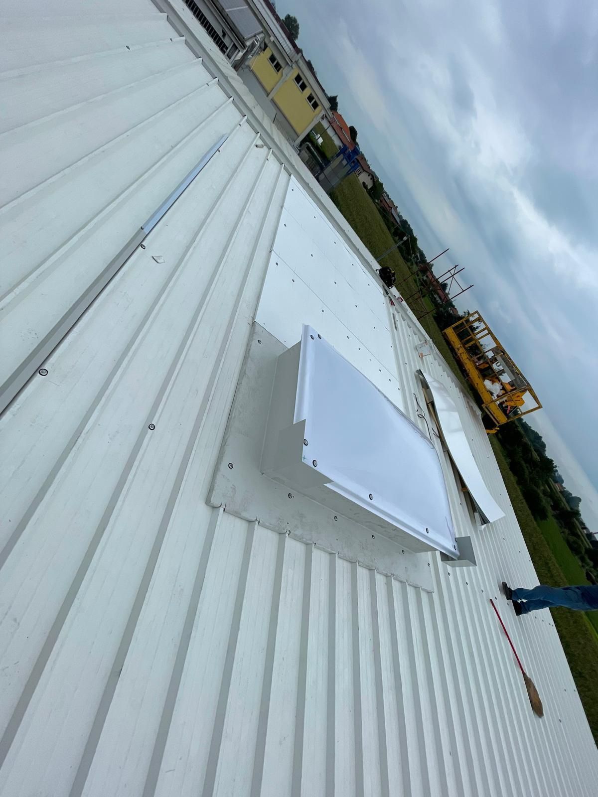 Tetto con pannelli ondulati bianchi e un piccolo lucernario sotto un cielo nuvoloso, con attrezzature da cantiere nelle vicinanze.