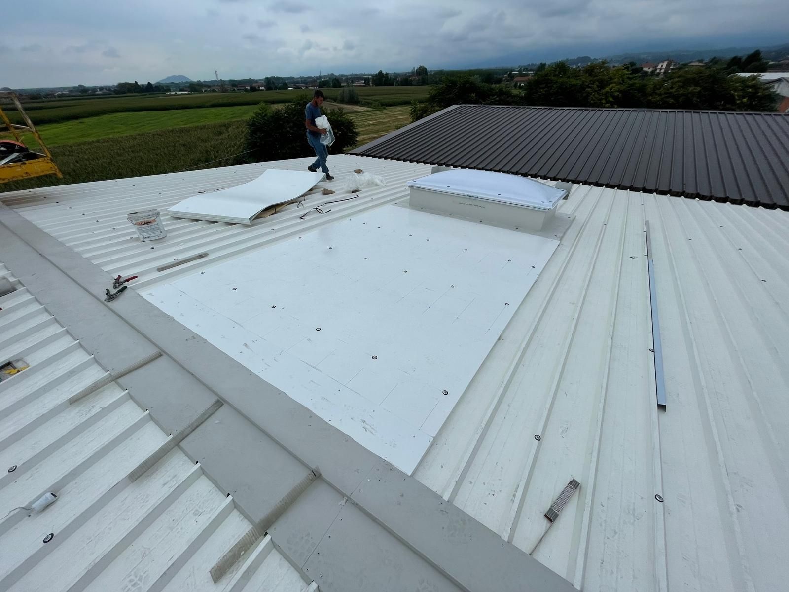 Un operaio su un tetto di metallo bianco ispeziona una sezione danneggiata vicino a un tetto scuro sotto un cielo nuvoloso.