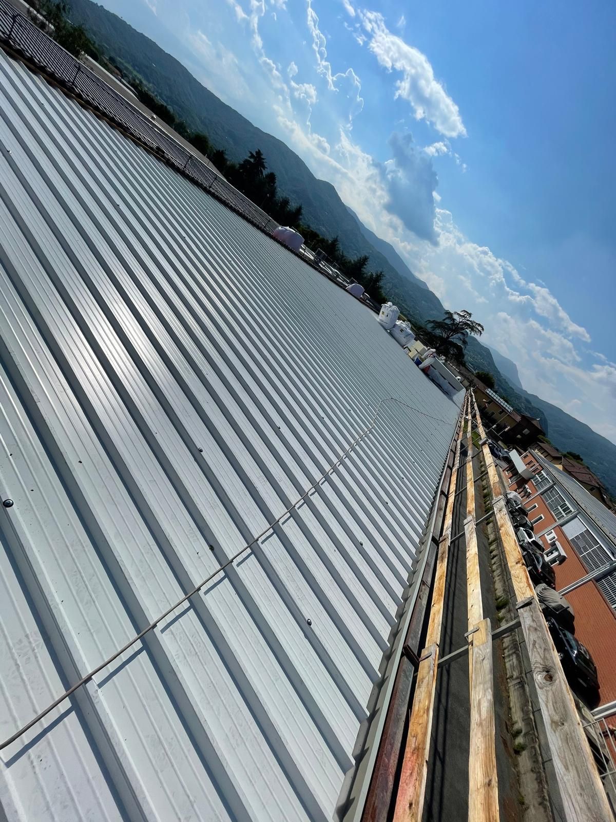 Vista obliqua di un tetto con pannelli metallici argentati e materiali da costruzione sotto un cielo azzurro.