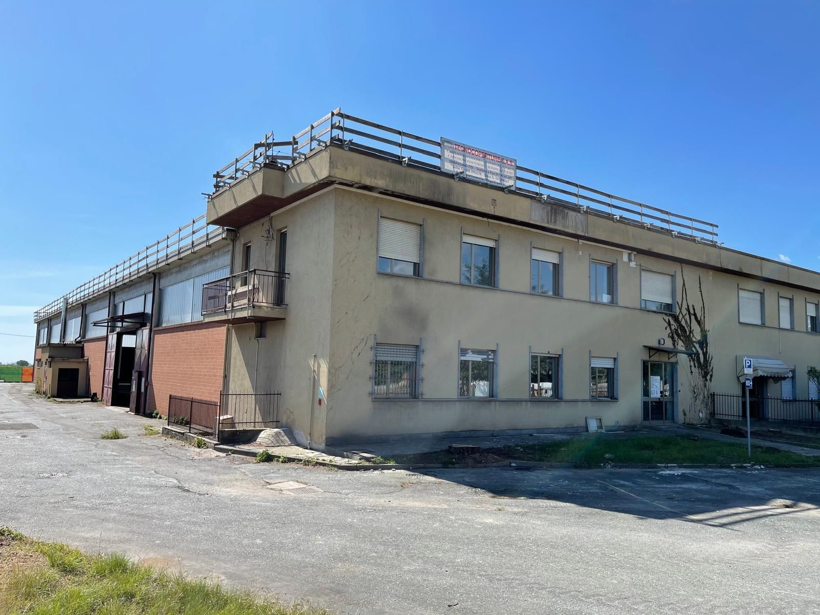 Edificio industriale di colore beige basso con ringhiere sul tetto e piazzale ghiaioso sotto un cielo azzurro limpido.