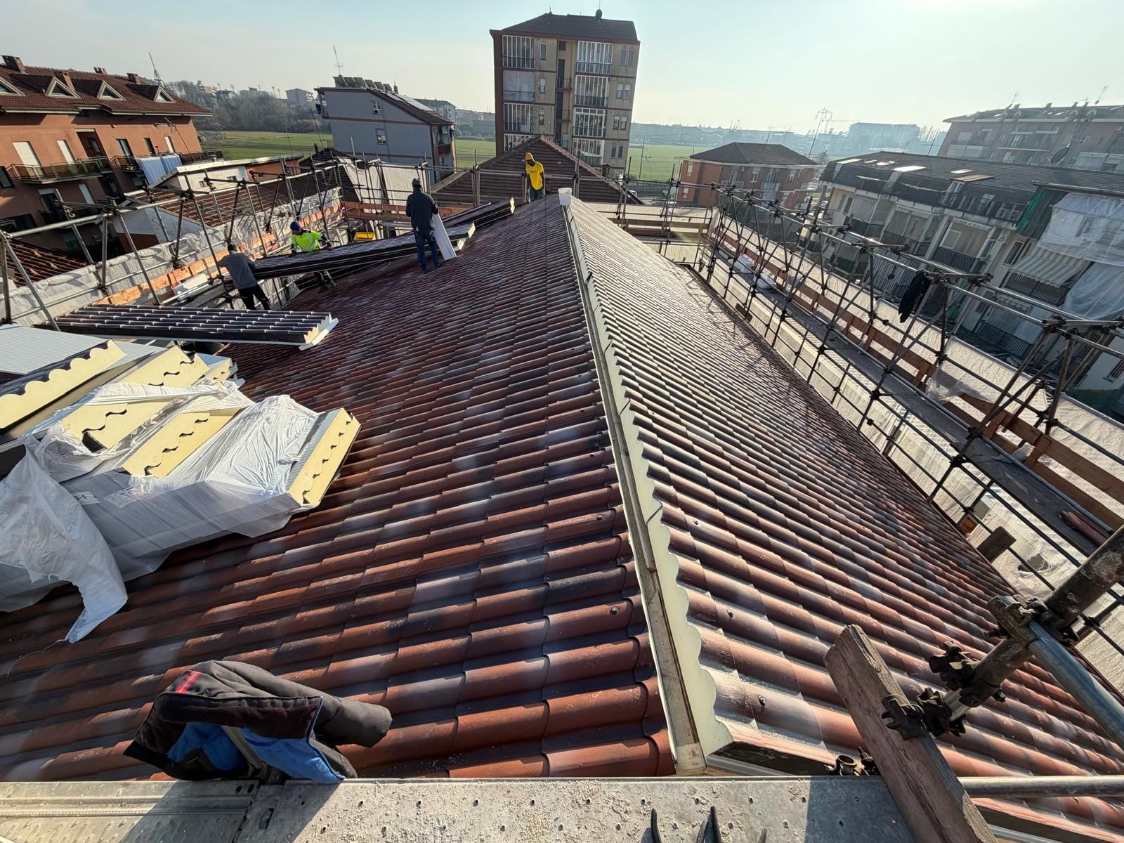 Operai al lavoro su un tetto di tegole rosse, accanto a un'impalcatura, con lo skyline della città sullo sfondo.