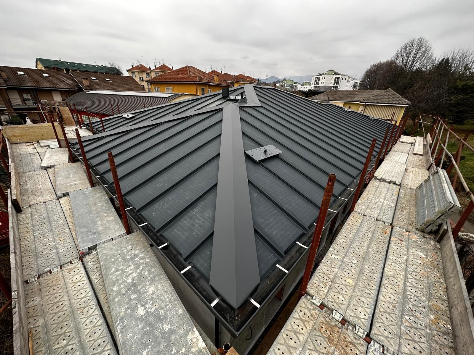 Tetto in costruzione con pannelli metallici a giunzione verticale di colore grigio scuro e impalcature su entrambi i lati.