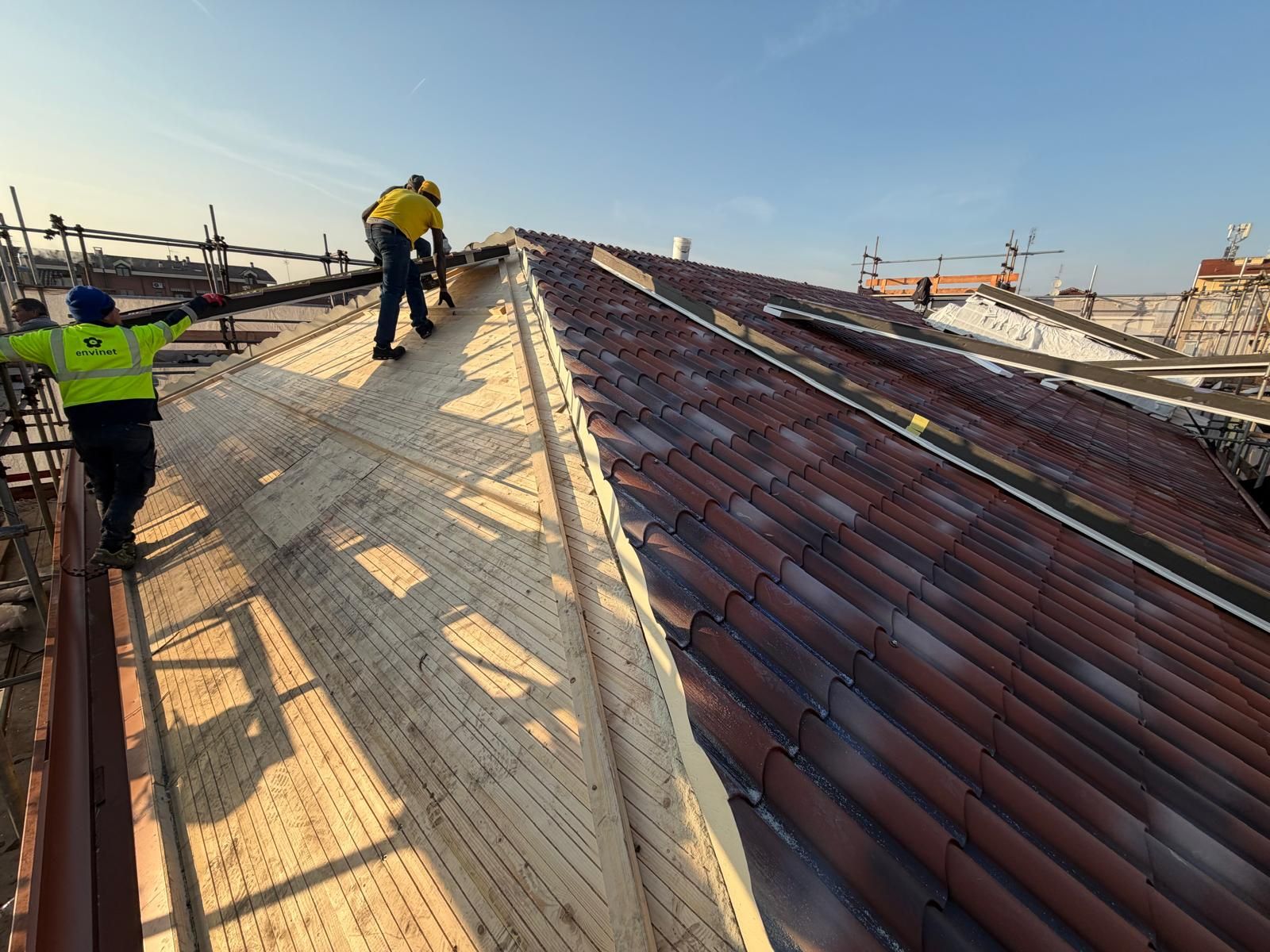 Operai intenti a riparare un tetto spiovente di tegole sotto la luce diretta del sole, con impalcature e tetti di edifici sullo sfondo.