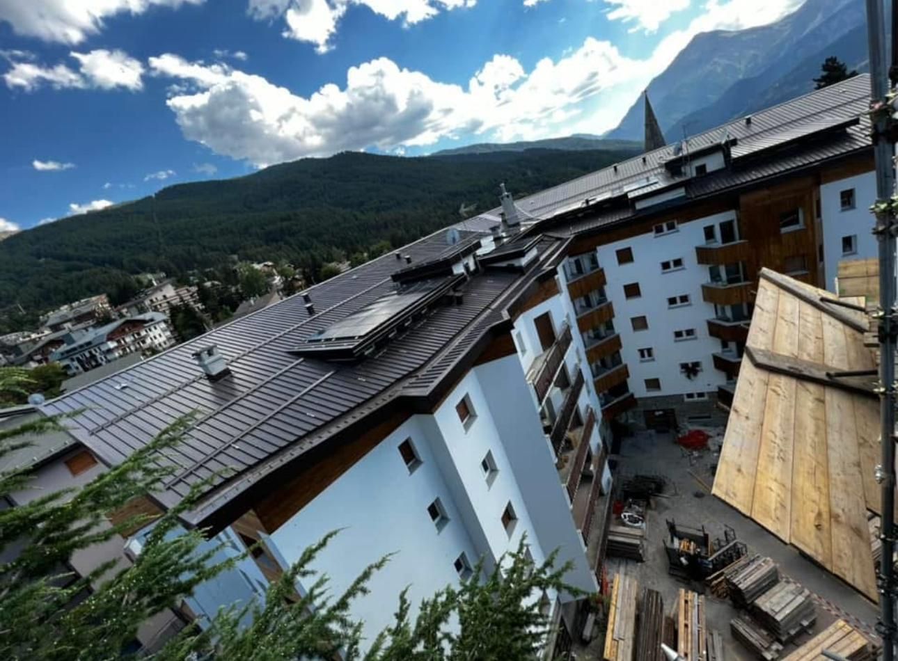 Veduta inclinata di edifici alpini bianchi con tetti scuri sotto un cielo azzurro e montagne sullo sfondo.