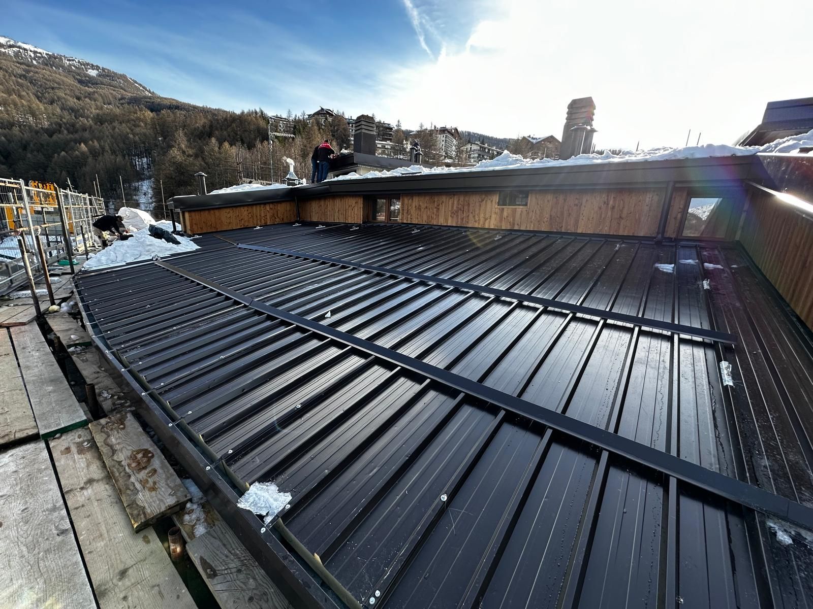 Un tetto di metallo nero ricoperto di neve accanto a un edificio in legno, sotto un cielo luminoso, con le colline sullo sfondo.