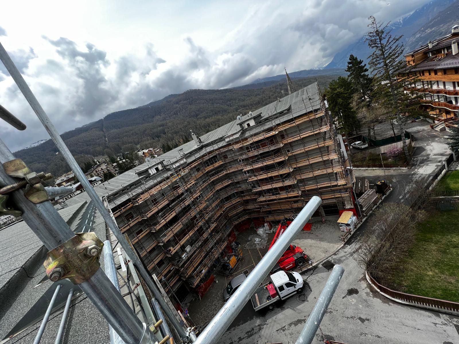 Edificio curvo a più piani in costruzione in una valle di montagna, con gru e camion nelle vicinanze.