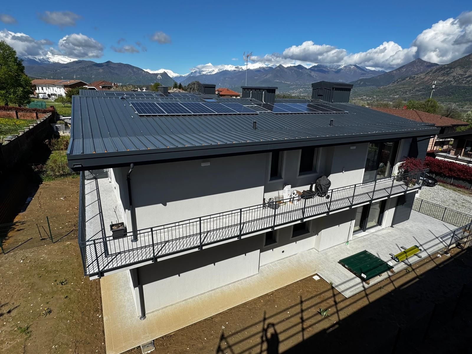 Casa a due piani con tetto scuro, balcone e sfondo montuoso sotto un cielo parzialmente nuvoloso.