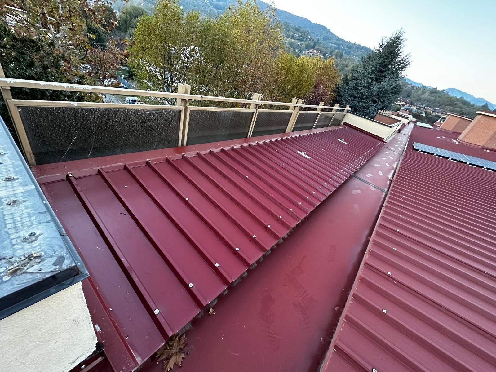 Tetto in metallo rosso con pannelli di colmo accanto a una recinzione, con alberi e montagne sullo sfondo.