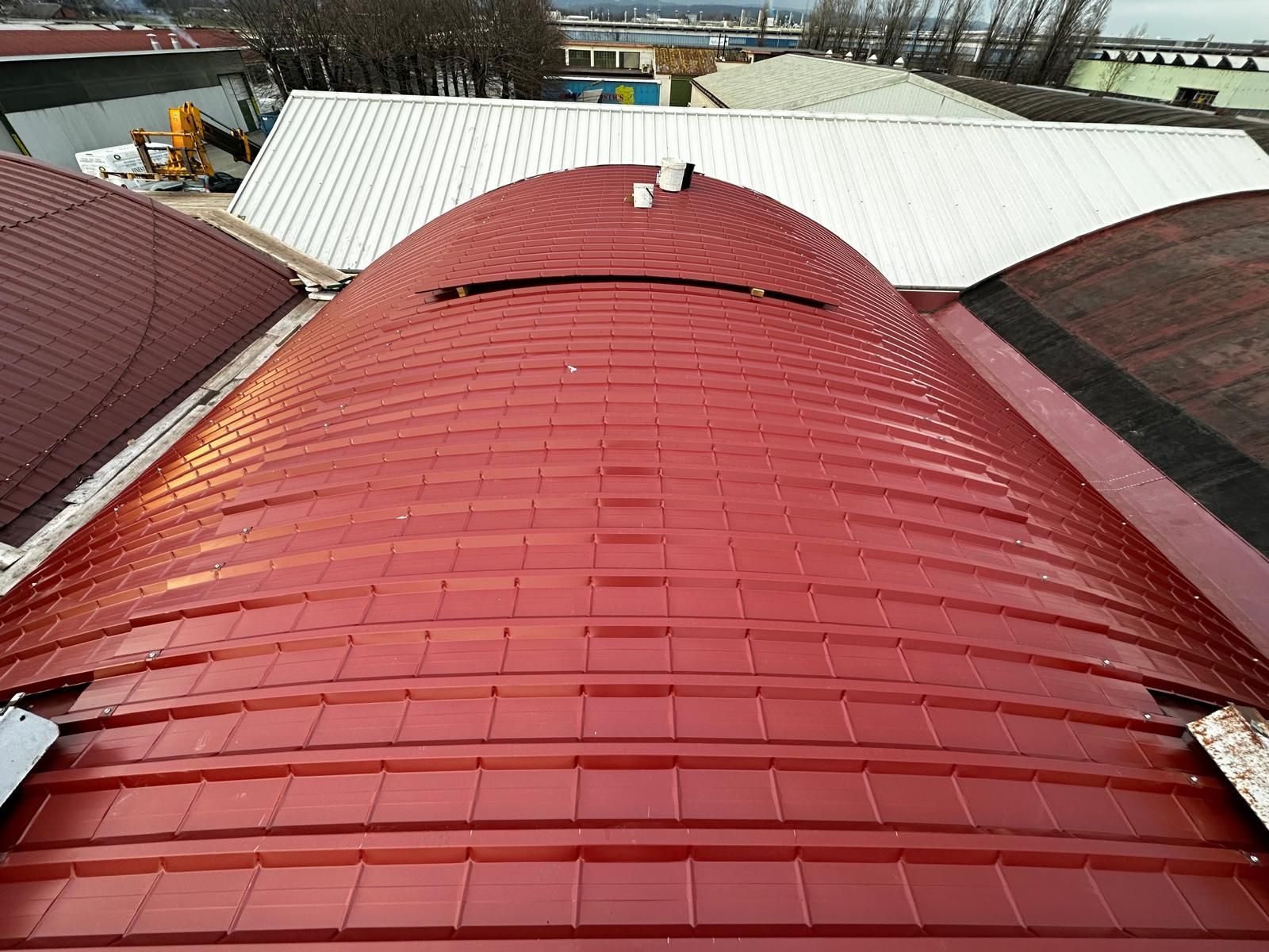 Tetto metallico rosso e curvo tra tetti bianchi e scuri, visto dall'alto.