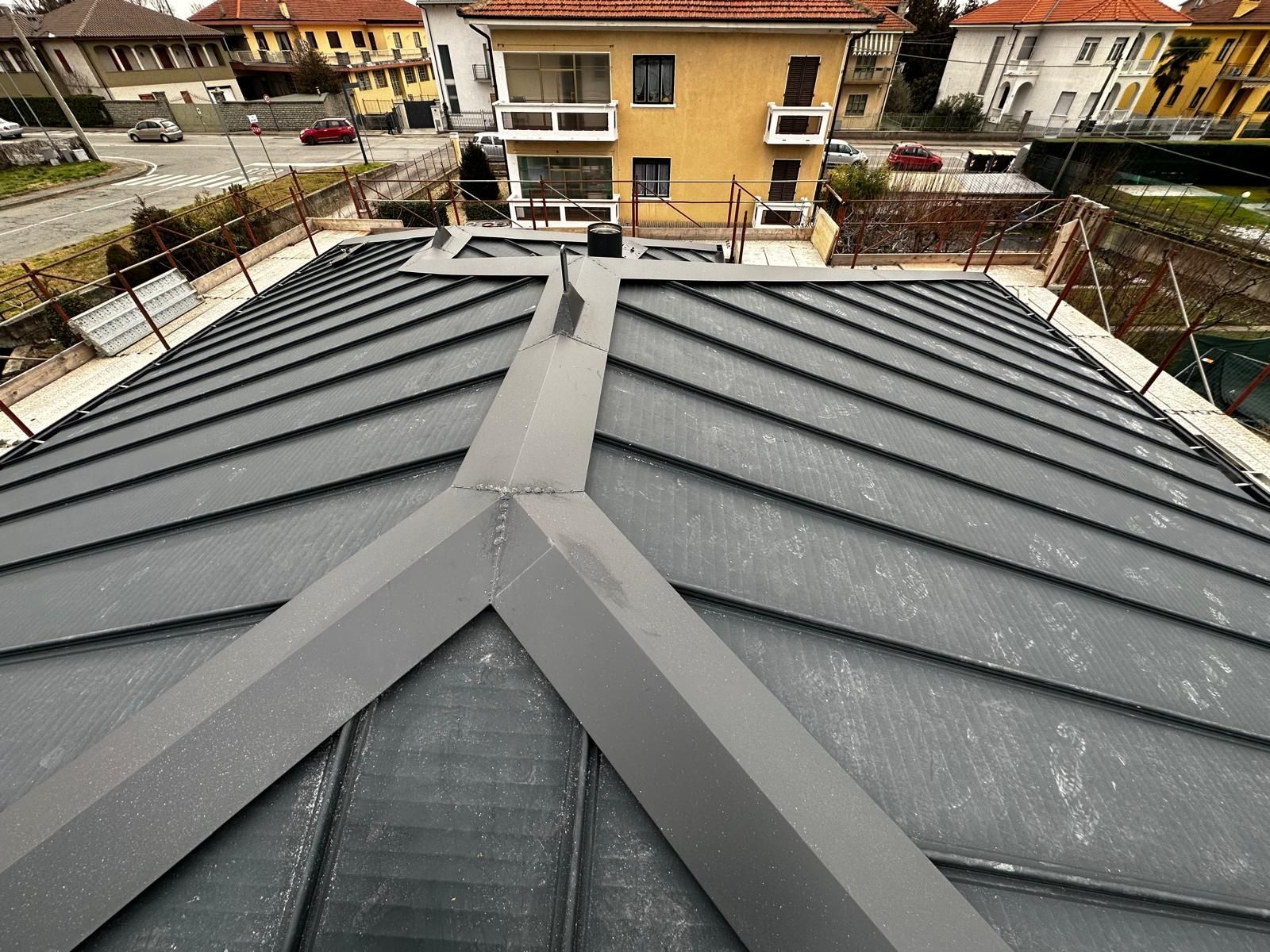 Tetto in metallo grigio con giunti verticali visto dall'alto, di fronte a case residenziali sullo sfondo.