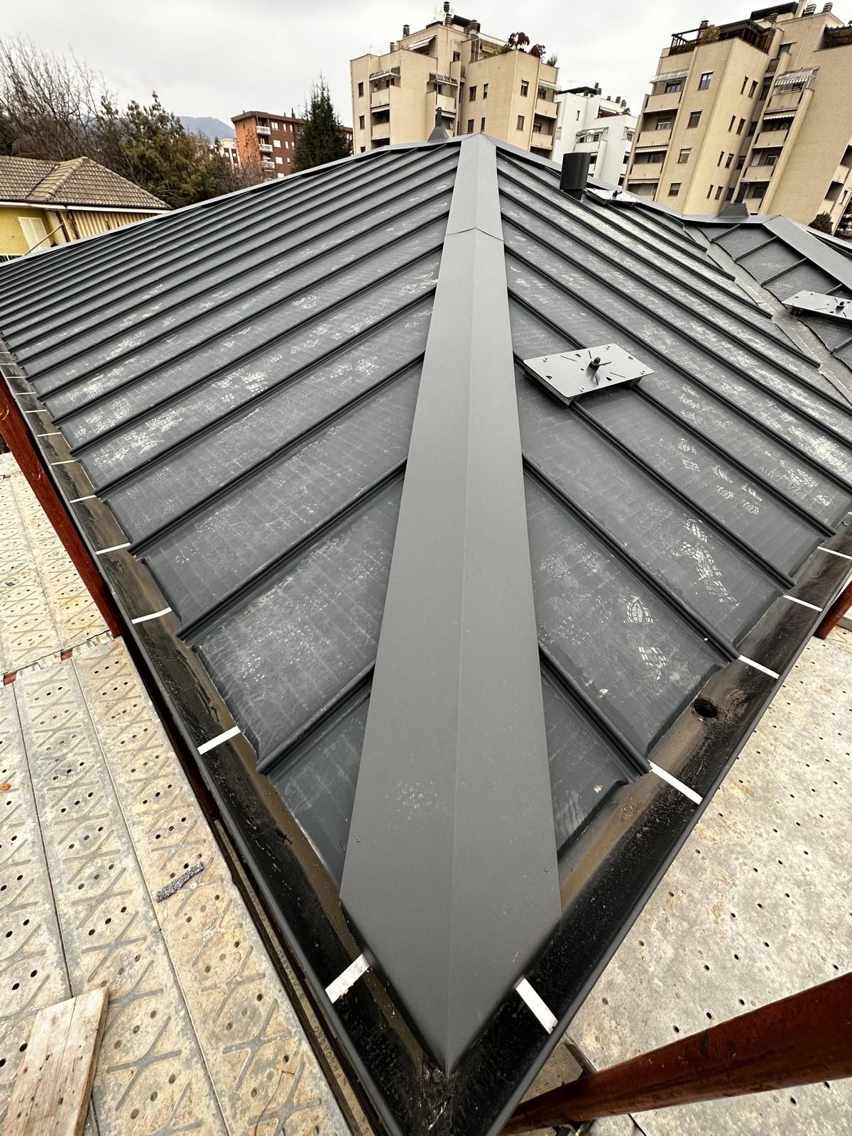 Tetto in metallo grigio con ventilazione centrale sul colmo, visto dall'alto, con palazzi residenziali sullo sfondo.