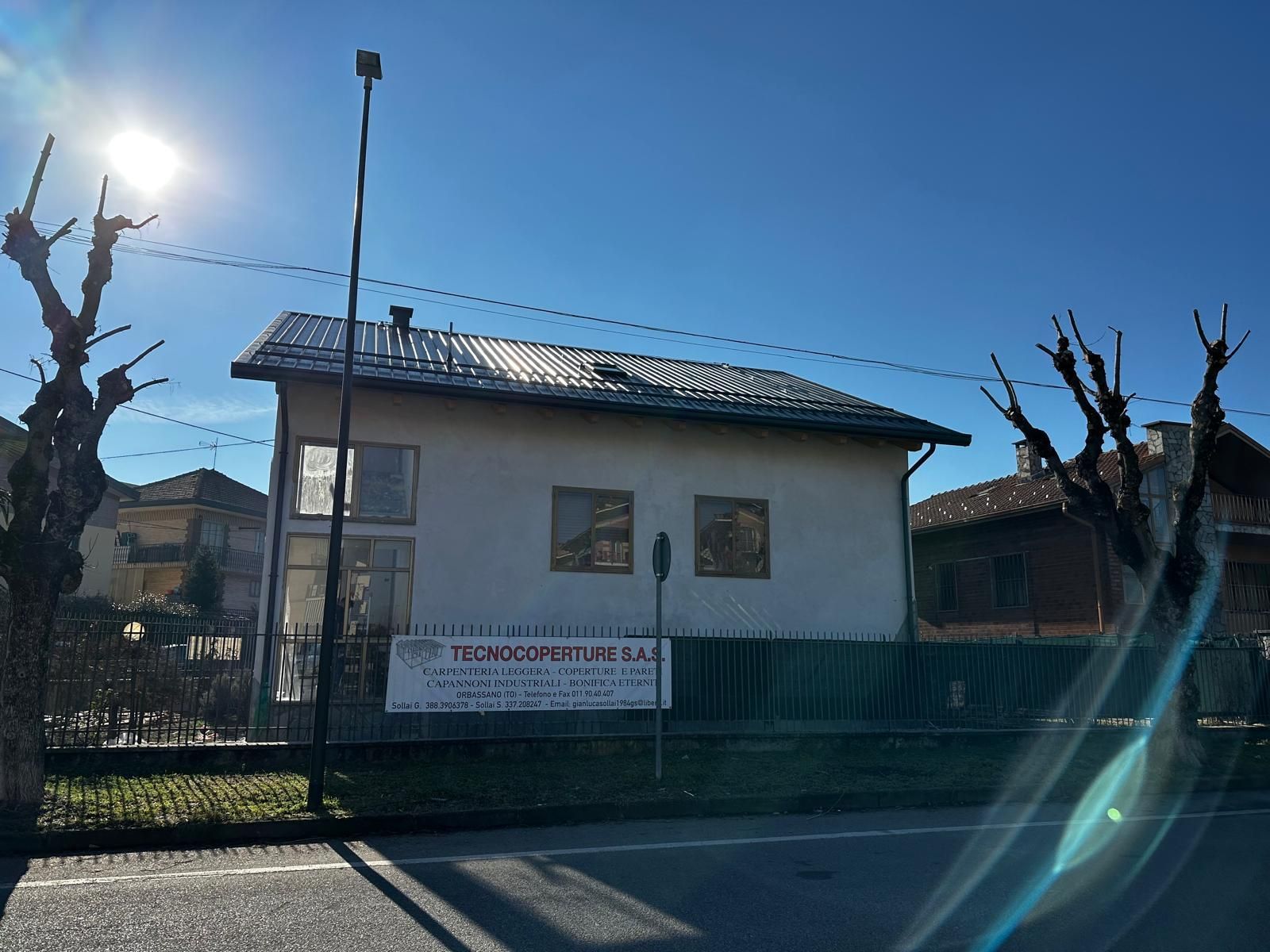 Un piccolo edificio bianco dietro una recinzione, sotto un cielo azzurro e soleggiato, con alberi spogli e uno striscione sulla facciata.