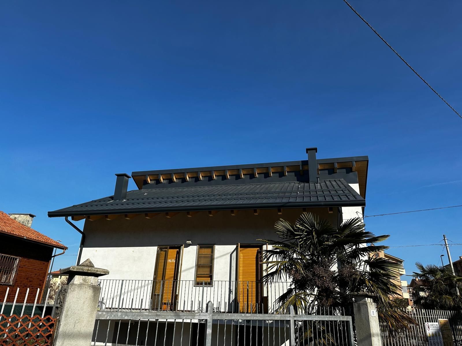 Casa bianca con tetto scuro e persiane di legno dietro una recinzione, sotto un cielo azzurro e limpido.