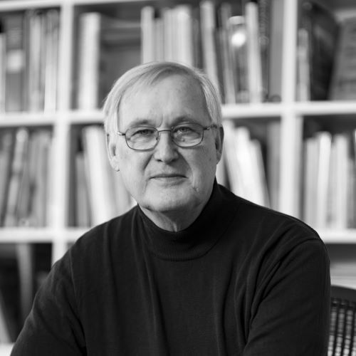 A man wearing glasses is sitting in front of a bookshelf in a black and white photo.