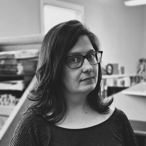 A woman wearing glasses and a black shirt is looking at the camera in a black and white photo.
