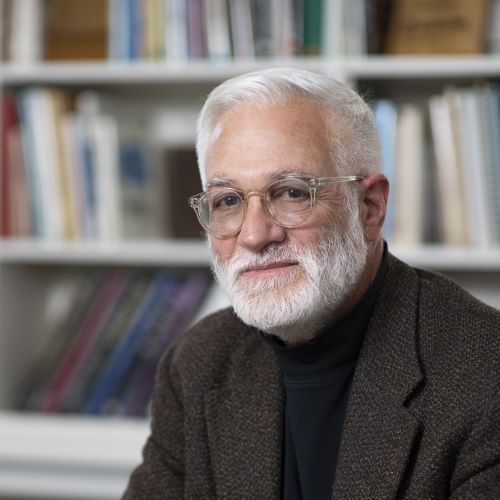 A man with a beard and glasses is sitting in front of a bookshelf.