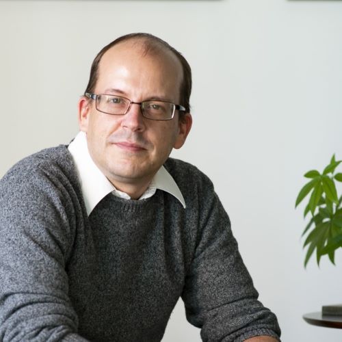 A man wearing glasses and a grey sweater looks at the camera