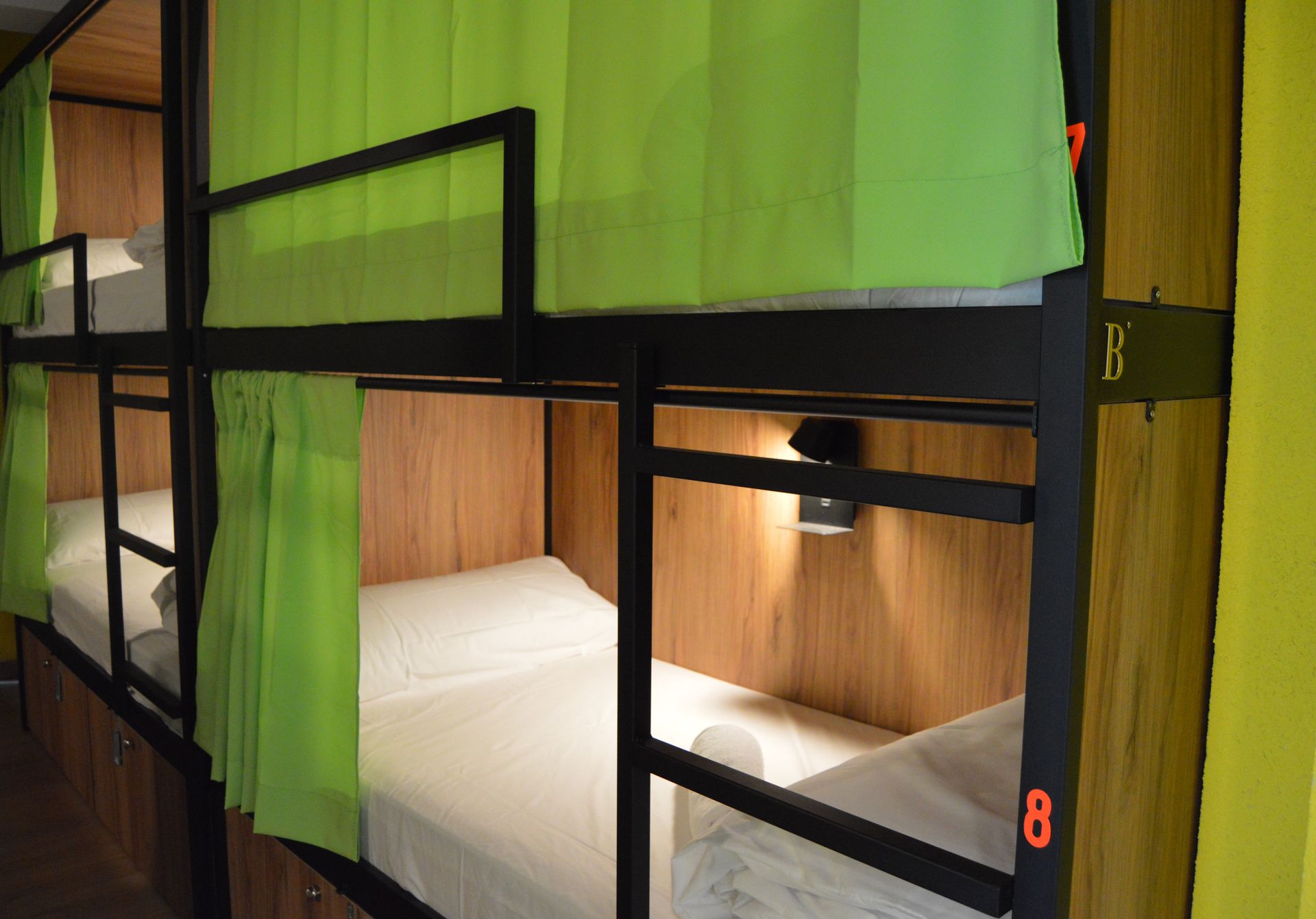 Literas con ropa de cama blanca, cortinas verdes y marcos negros en un albergue.