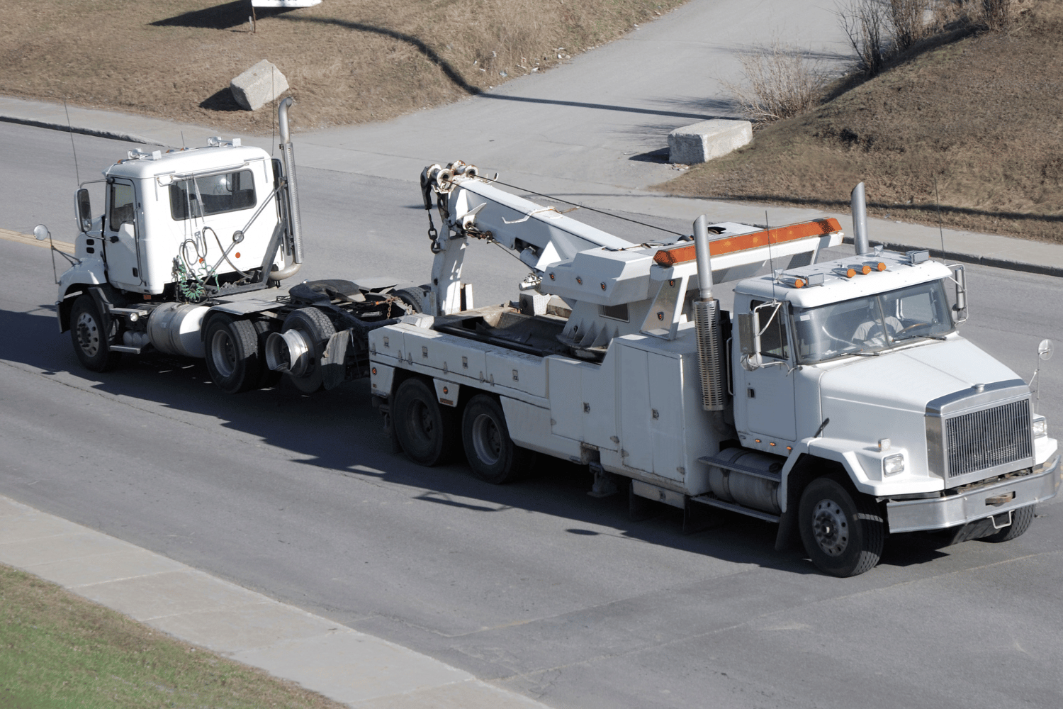 Two white tow trucks parked on a road, one hauling the other with a raised boom.