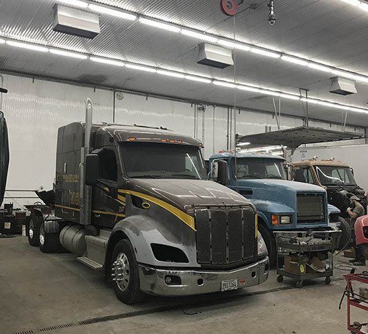 Three semi trucks parked inside a repair shop, including a gray and black truck in front.