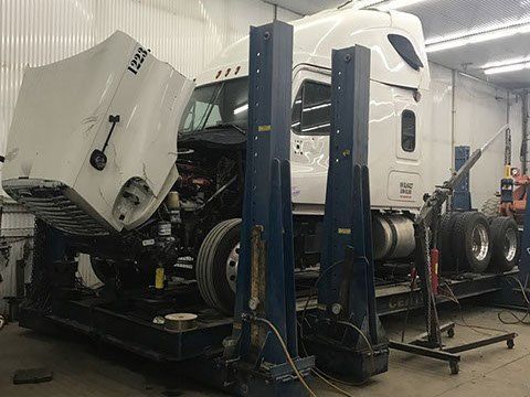 White semi-truck in a repair shop, front cab lifted for maintenance on a blue hydraulic lift.