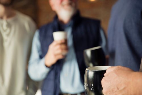 People holding mugs, blurred background, dark mugs, person in vest with mug.