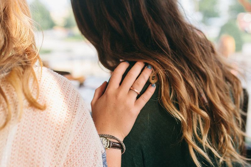 Person's hand on another's shoulder, offering support.  One has long wavy brown hair, the other has blonde hair, outdoors.