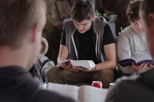 People seated, reading books. Man in center wears gray vest, focused. Others read, study.