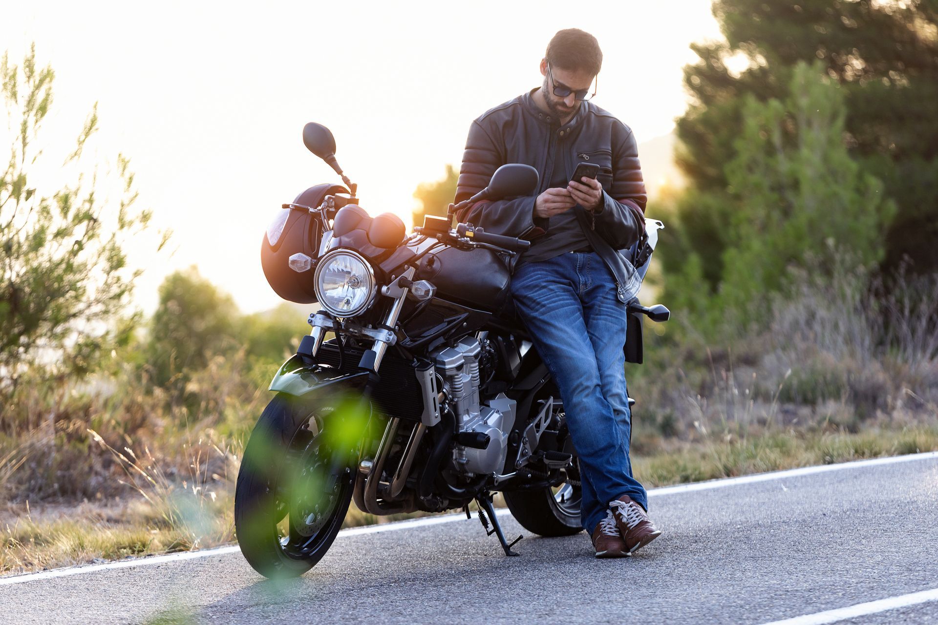 Man leaning on a black motorcycle on a rural road at sunset, looking at his phone.