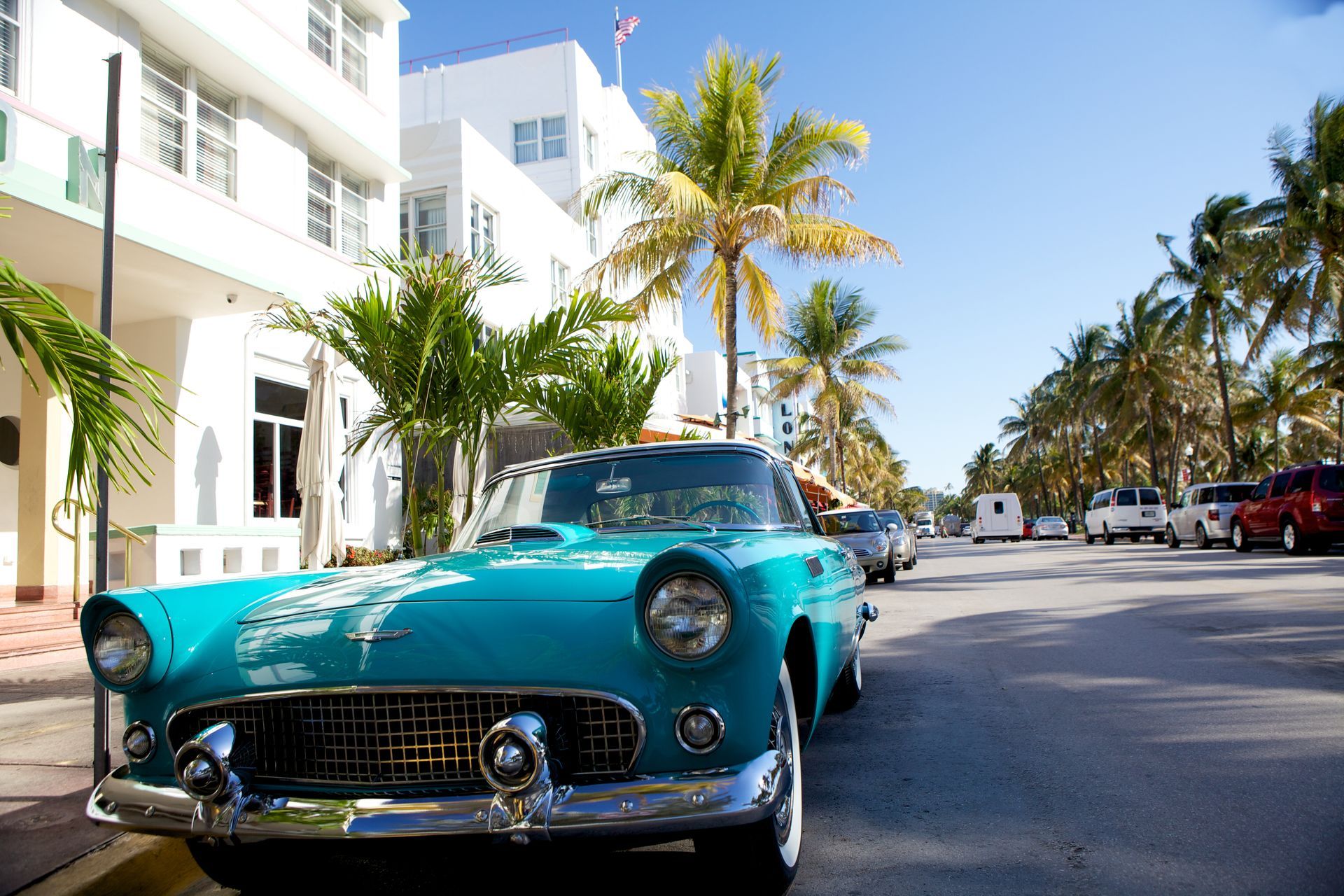 Turquoise vintage car parked on a sunny palm-lined street with white buildings in the background