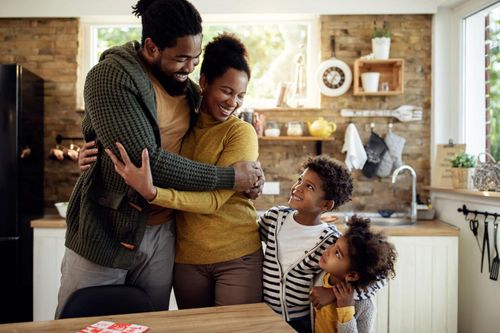 Family hugging in a bright kitchen, smiling around a table with two children and a baby in a carrier