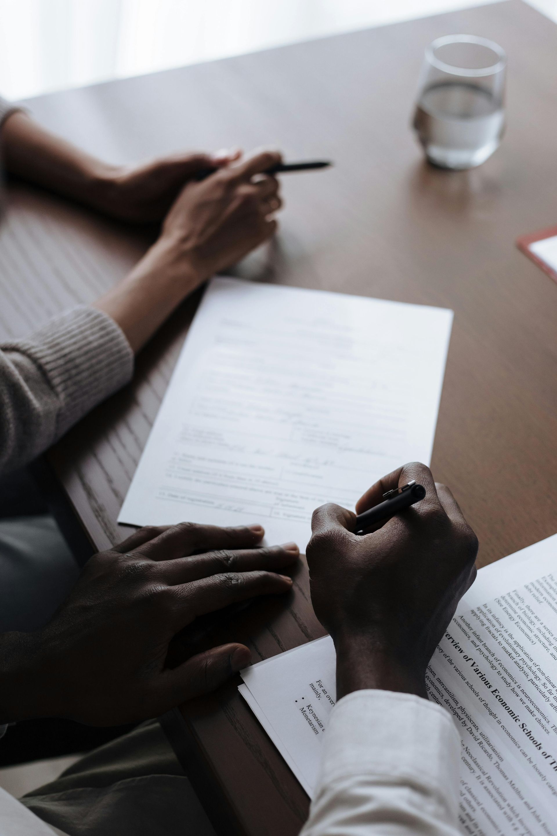 Two people writing notes at a table with papers and a glass of water