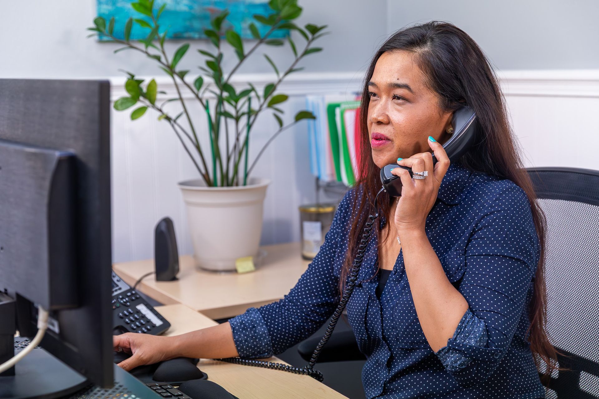 Woman in blue blouse talking on a phone at a desk with a computer and potted plant in the office
