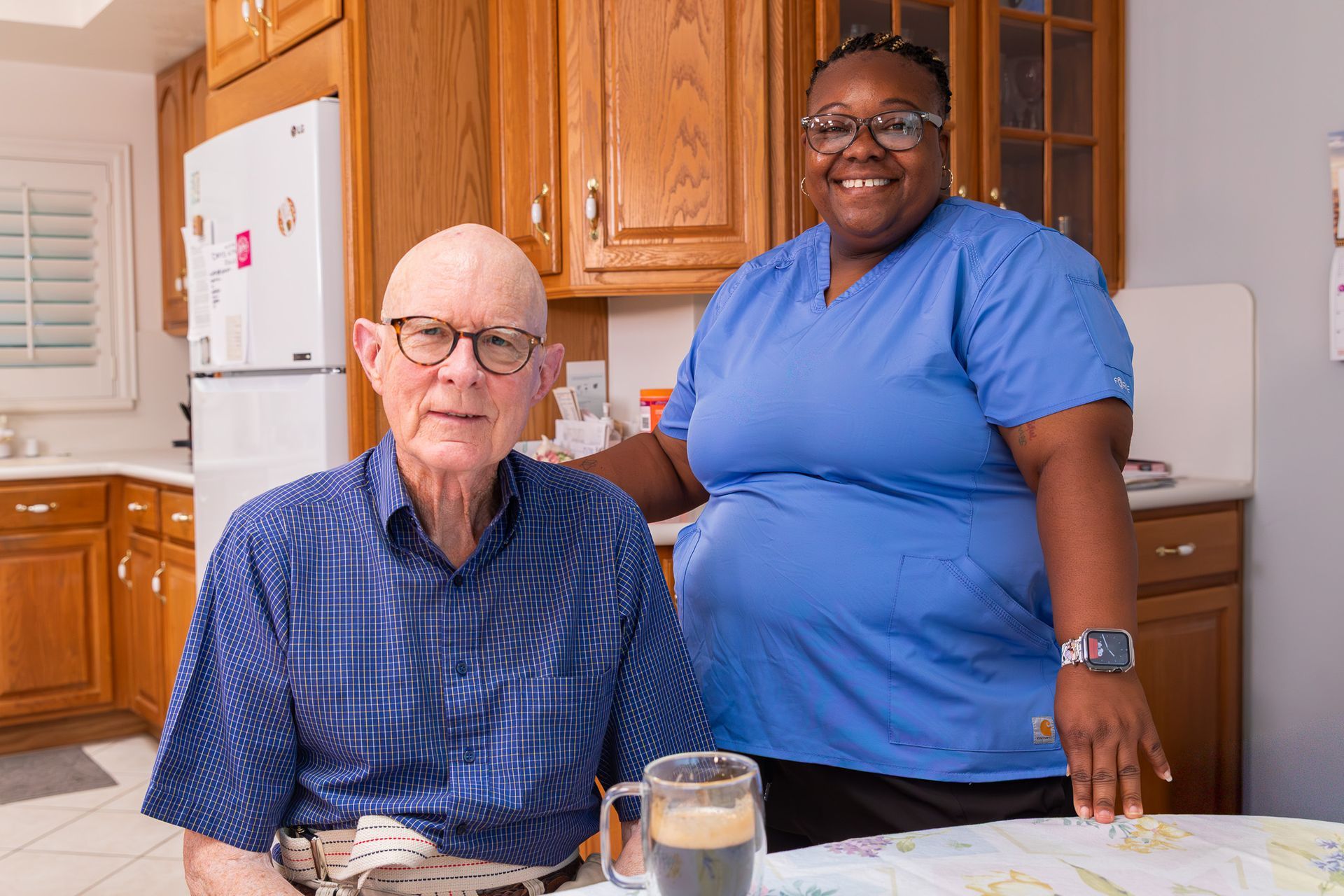 Two people smiling in a kitchen, standing beside a seated man at a table with breakfast items.